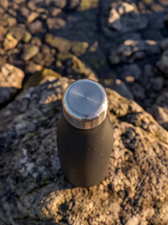 A B3TTER bottle resting on rough rock at the edge of the water